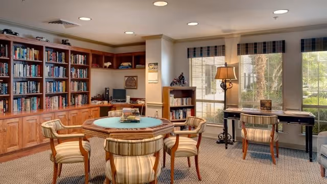 A cozy senior living common area with a round wooden table surrounded by four striped upholstered chairs. Behind the table is a large wooden bookshelf filled with books and decorative items. To the right, there is a window with blinds and a small desk with two chairs, a lamp, and some books. The room has carpeted flooring and soft lighting.