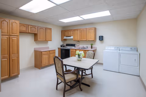 Small institutional kitchenette and laundry area with wooden cabinets, a stove, washer and dryer, and a table with chairs.