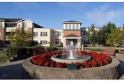 A circular fountain surrounded by red flowers and benches sits in front of the entrance to a multi-story senior living building under a clear blue sky.