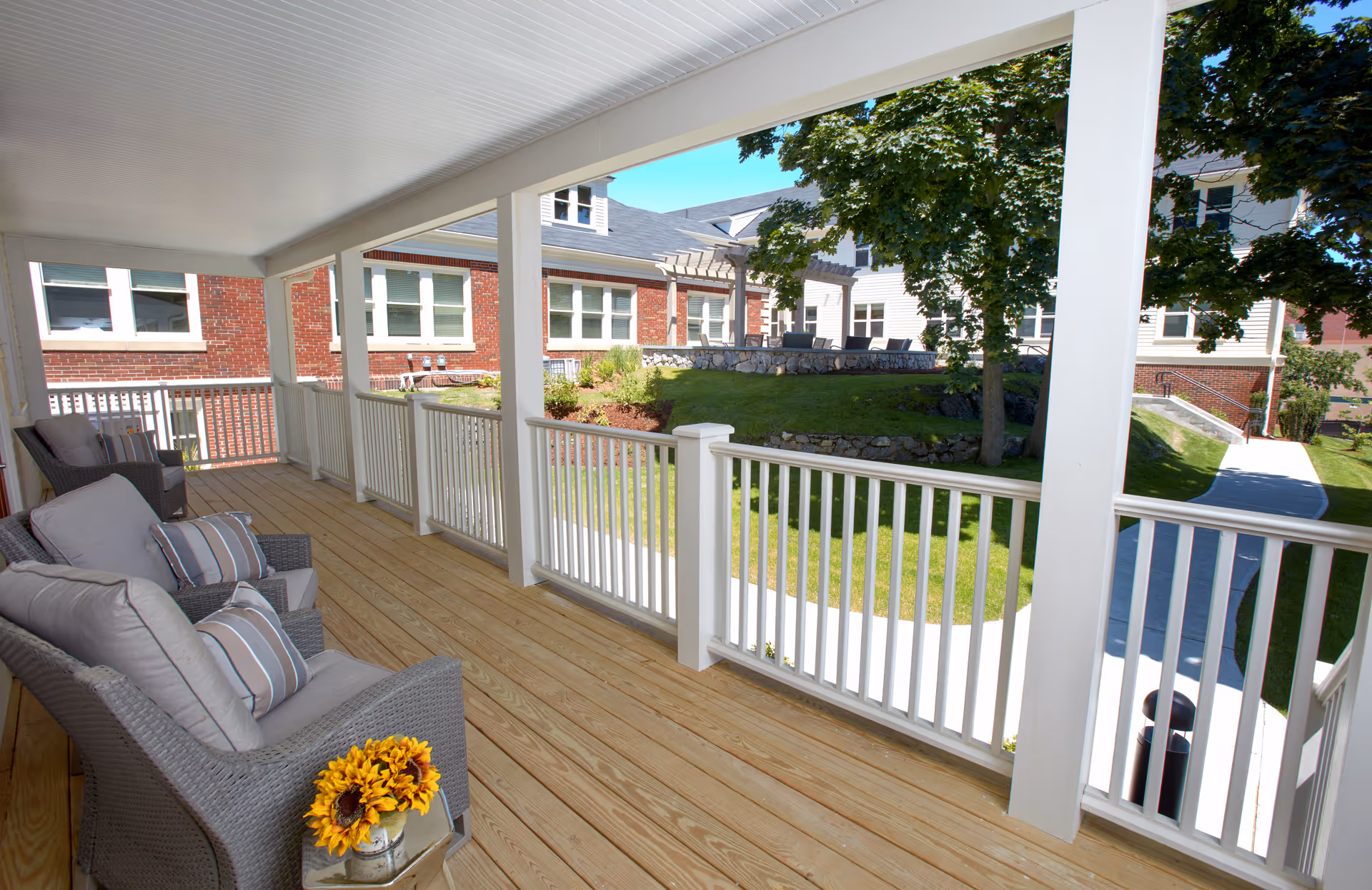Covered wooden porch with cushioned wicker chairs and a small table overlooking a landscaped courtyard and brick senior living buildings.