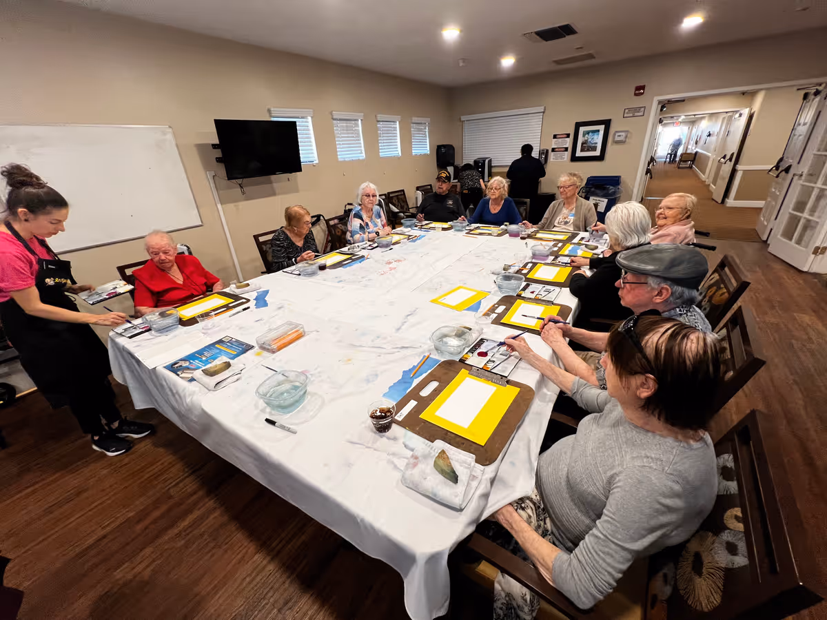 A group of elderly individuals seated around a large rectangular table covered with a white tablecloth, participating in a painting activity with art supplies and water bowls in front of them. A staff member stands nearby assisting one of the participants. The room has beige walls, a mounted TV, and windows with blinds.