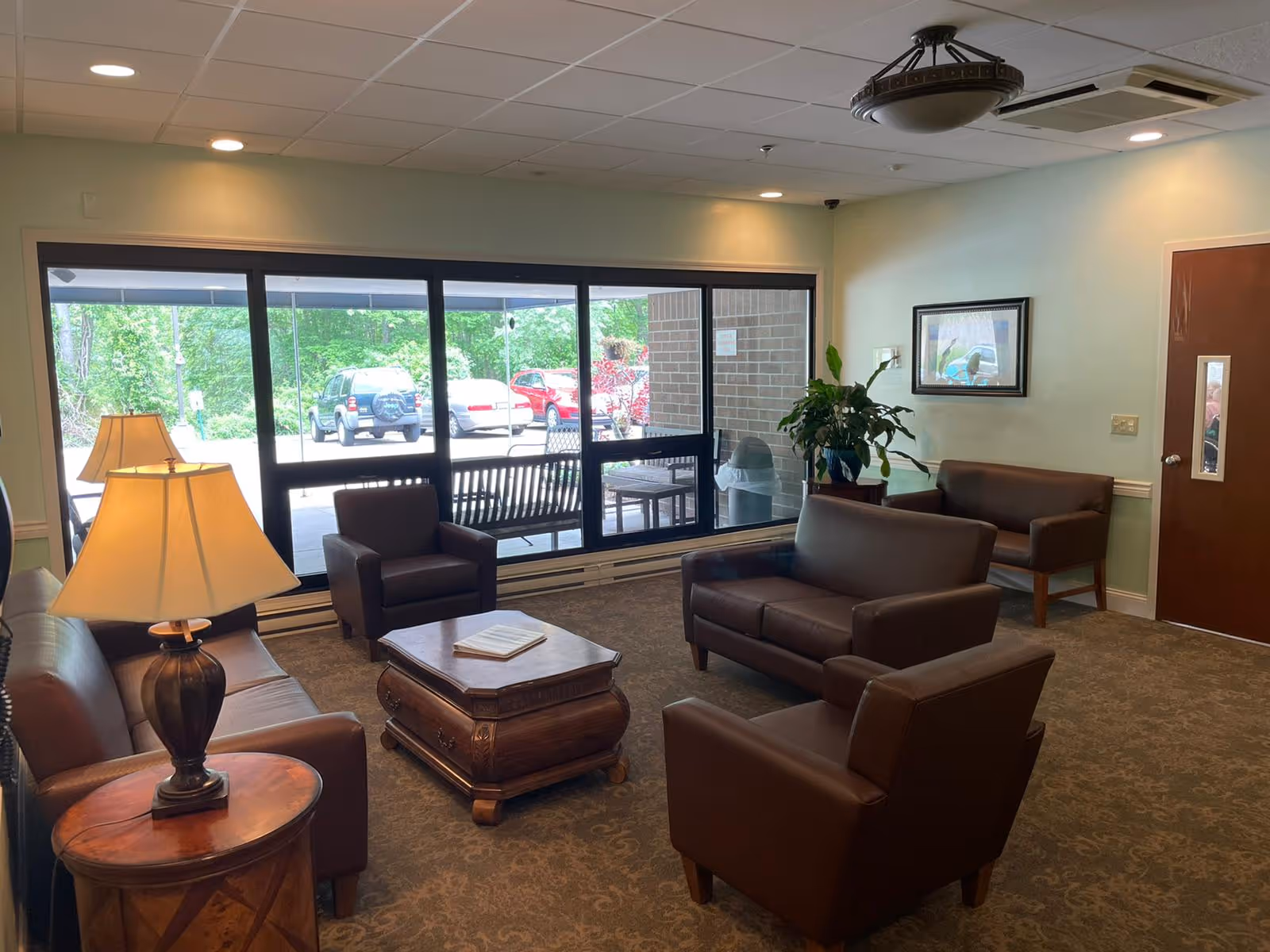 A cozy nursing home lounge area with brown leather sofas and armchairs arranged around a wooden coffee table. There is a side table with a lamp, a potted plant on a small table, a framed picture on the wall, and large windows showing a parking lot and greenery outside.