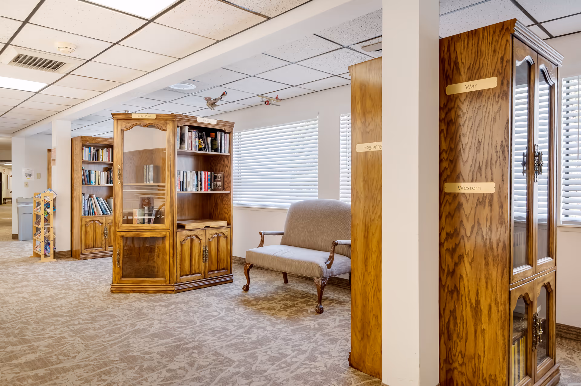 Carpeted interior reading area with wooden bookcases, a cushioned loveseat, and labeled shelves by windows.