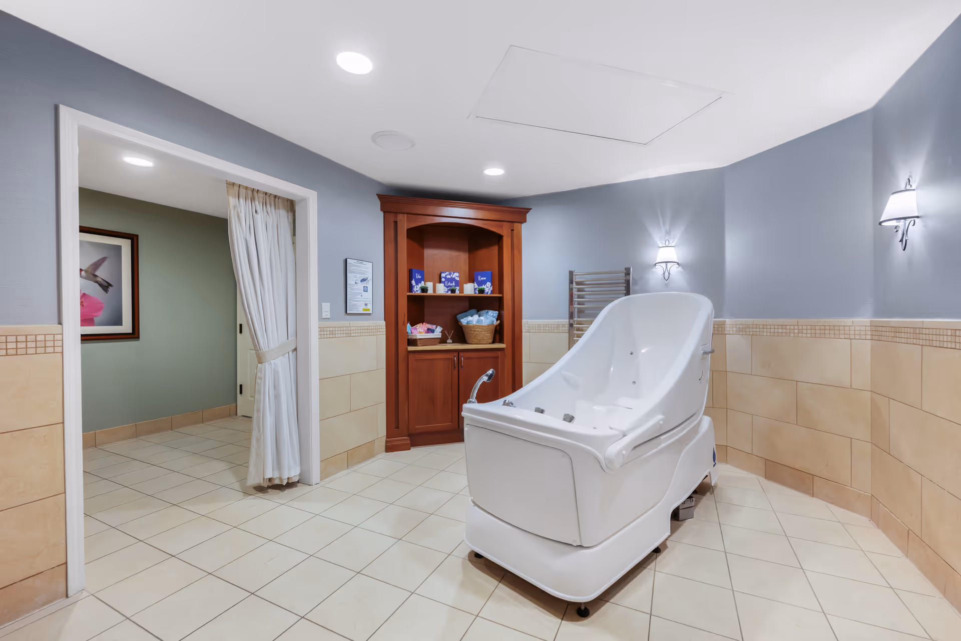 A clean, well-lit bathing room with beige tiled walls and floor. The room features a large white assisted bathing tub in the center, two wall-mounted light fixtures, a wooden cabinet with shelves holding bath supplies, and an open doorway with a curtain leading to another room with green walls and a framed picture of a hummingbird.