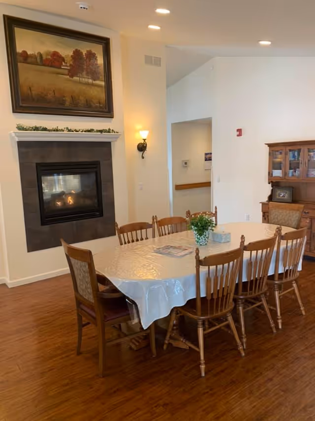 A dining room with a rectangular table covered with a white plastic tablecloth surrounded by eight wooden chairs. On the table, there is a small green plant and a tissue box. The room has wooden flooring, a wall-mounted fireplace with a painting of a landscape above it, and a wooden cabinet with glass doors in the background. The walls are painted white, and there is a wall sconce light fixture next to the fireplace.