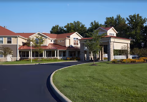 Exterior view of a senior living facility with a curved driveway, green lawn, and a covered entrance. The building has beige siding with red roofs and multiple windows, surrounded by trees and landscaping.