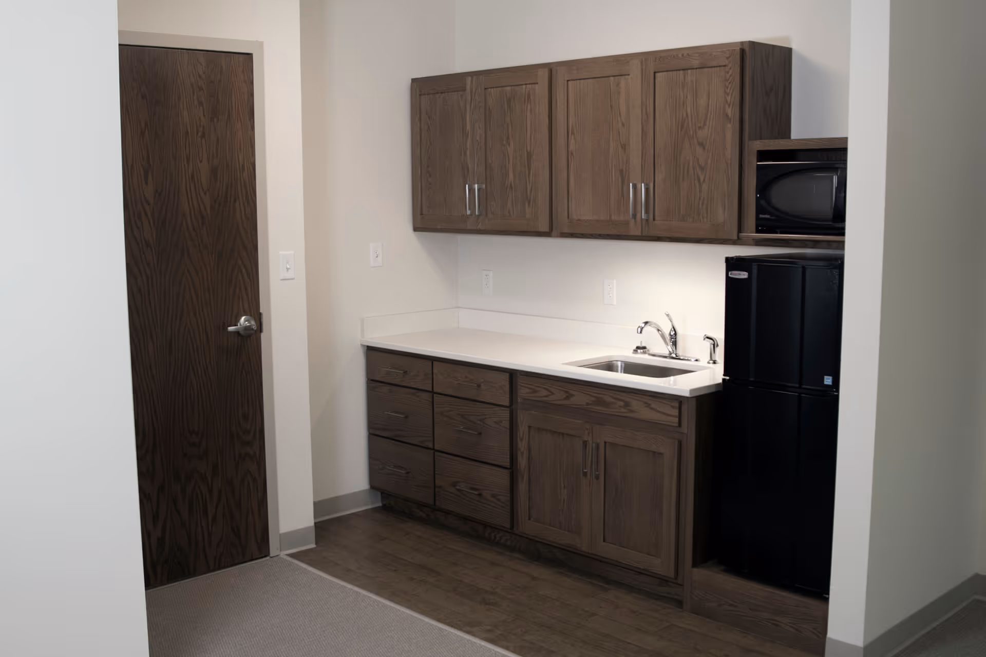 Small kitchenette with wood cabinets, a countertop and sink, a black mini-fridge and microwave beside a closed wooden door.