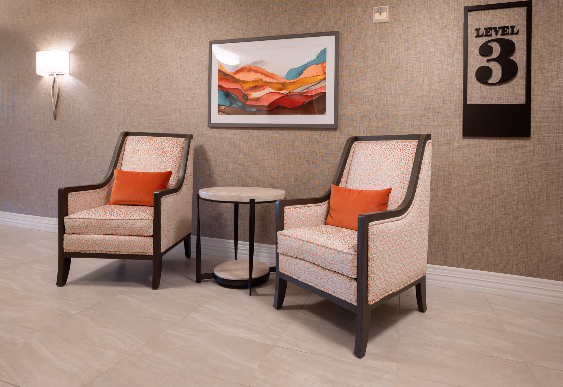 Two upholstered armchairs with orange accent pillows are positioned on either side of a small round table in a hallway or lounge area. The wall behind them is covered in textured wallpaper and features a colorful abstract painting and a sign that reads 'LEVEL 3'. A wall-mounted light fixture is visible on the left side.