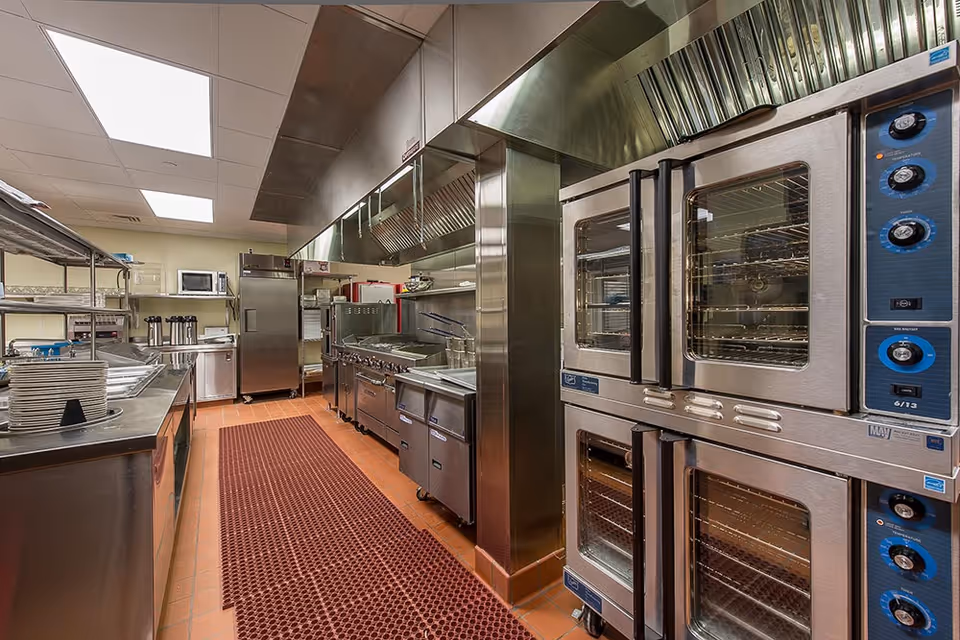 A clean and well-equipped commercial kitchen with stainless steel appliances including ovens, stovetops, a refrigerator, microwave, and metal shelving. The floor has a red rubber mat runner and the ceiling has bright fluorescent lighting.