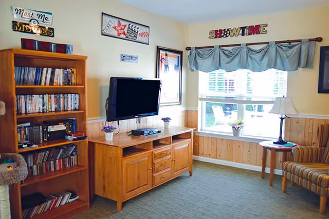 A cozy living room area with a wooden bookshelf filled with DVDs and books, a wall-mounted flat-screen TV above a wooden cabinet, a window with blue valance curtains, a small round table with a lamp, and a striped armchair. The walls are decorated with movie-themed signs and posters.