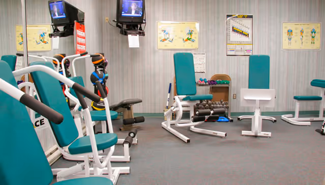 A small exercise room with several pieces of fitness equipment including seated weight machines and dumbbells. The walls have anatomical charts and two small televisions mounted near the ceiling. The floor is carpeted and the room has a calm, organized atmosphere.