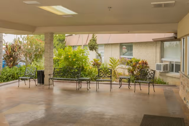 Covered outdoor seating area with metal benches and chairs arranged on a concrete floor, surrounded by greenery and plants near a building with windows and an air conditioning unit.