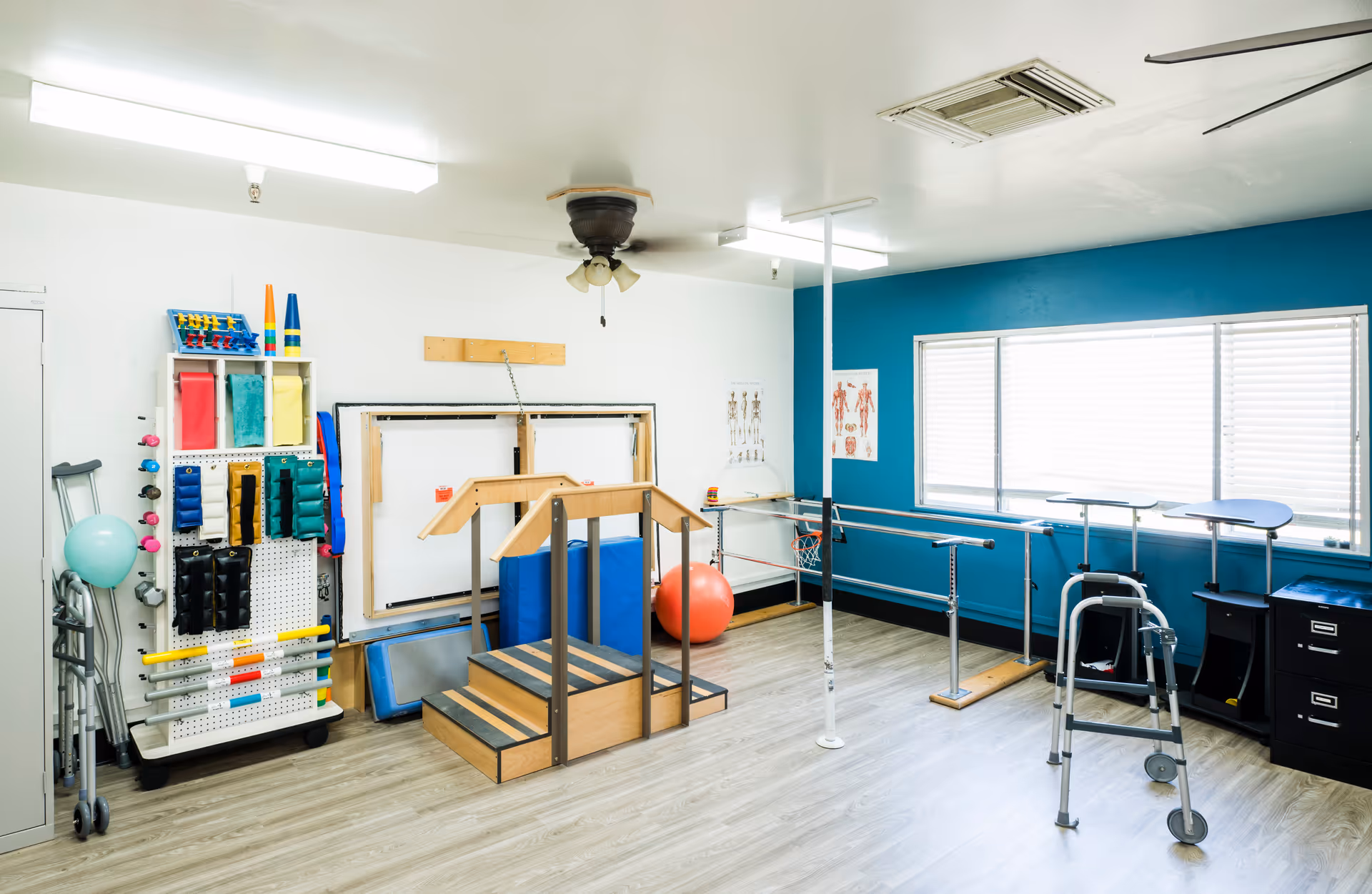 A bright physical therapy room with wooden steps, parallel bars, a walker, exercise balls, and various therapy equipment against white and blue walls with a large window.