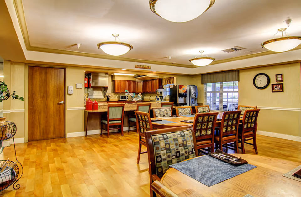 Well-lit communal dining area with wooden tables and chairs and an open kitchen counter in the background.