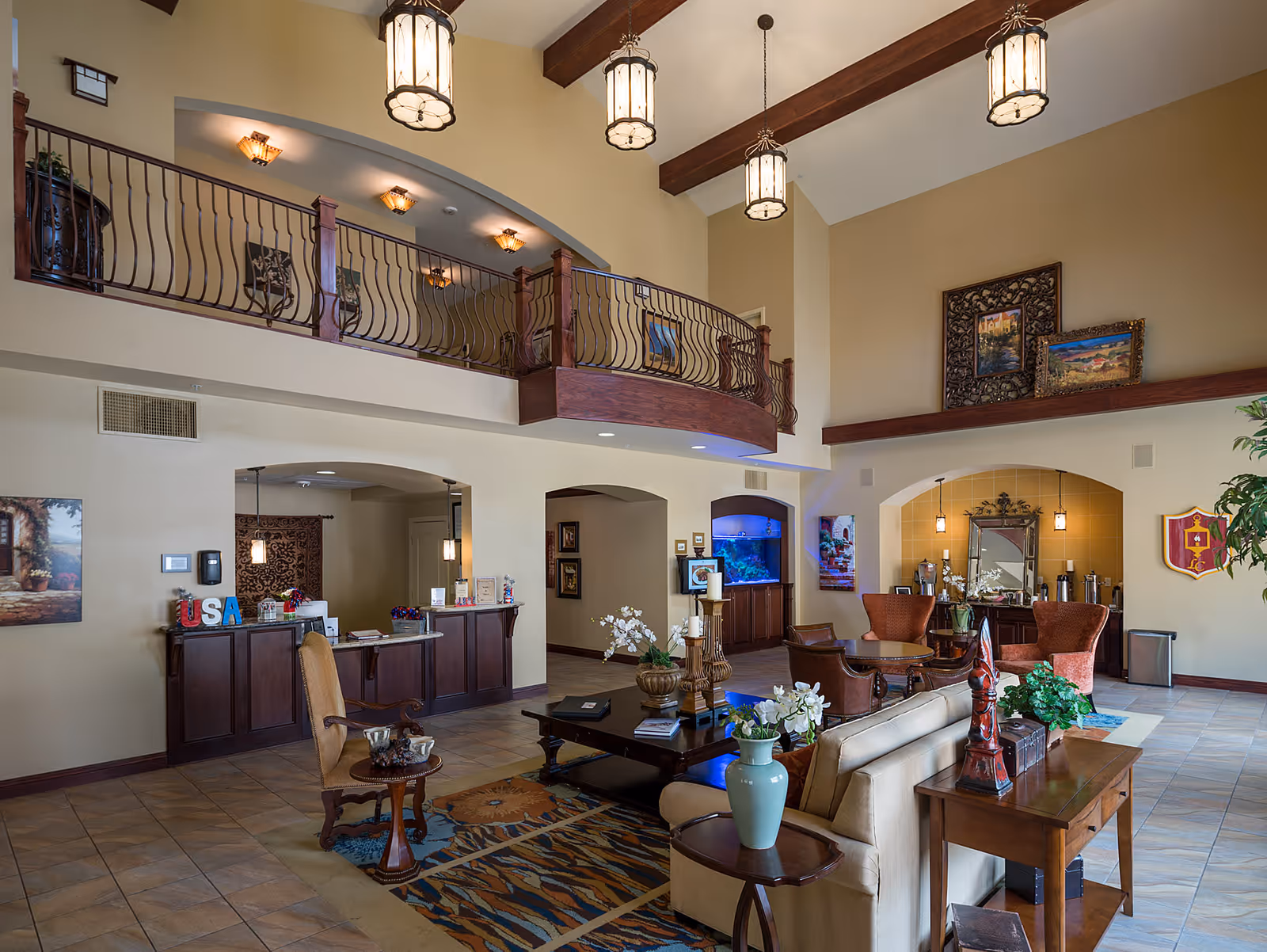 Spacious senior living facility common area with high ceilings, hanging lantern-style lights, a balcony with wrought iron railing, and a seating area featuring armchairs, a sofa, coffee tables, and decorative plants. There is a reception desk on the left side and an aquarium built into the wall in the background. The walls are adorned with framed artwork and decorative items.