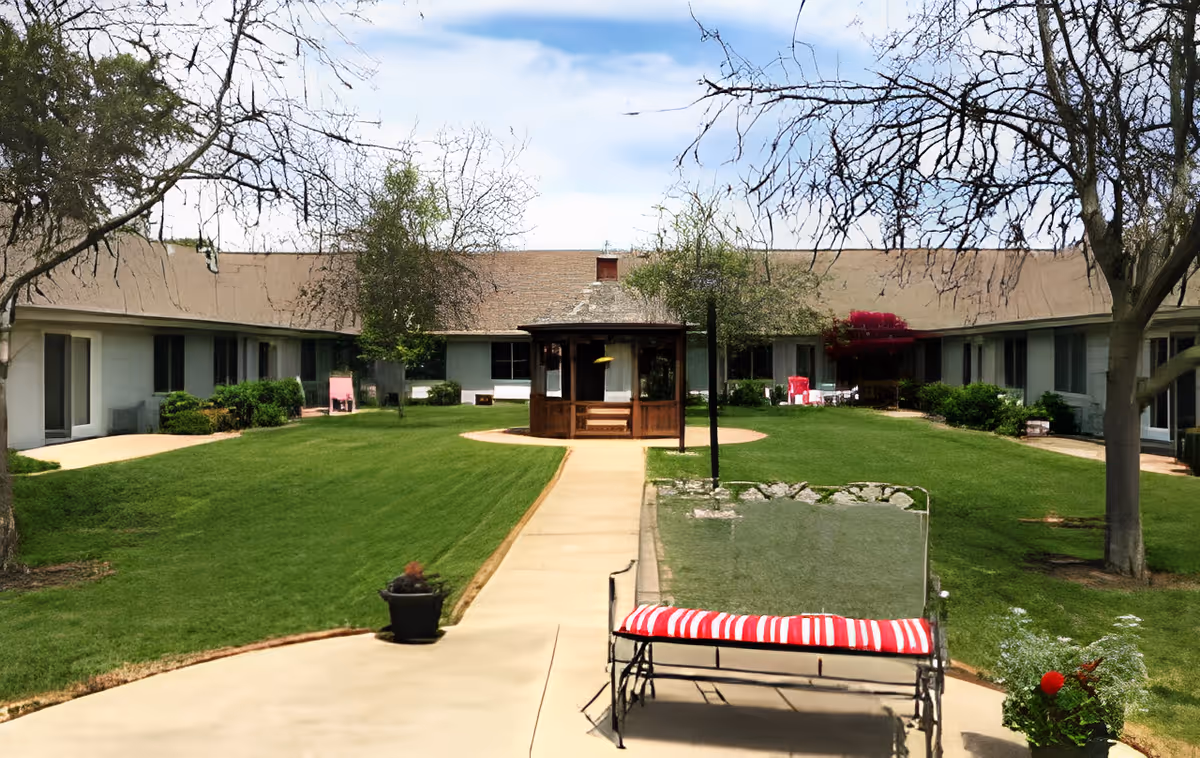 Outdoor courtyard area at Villas at Kewanee featuring a central wooden gazebo, surrounded by a well-maintained lawn, trees, and a curved building with multiple windows and doors. There is a paved walkway leading to the gazebo and a bench with a red and white striped cushion in the foreground.
