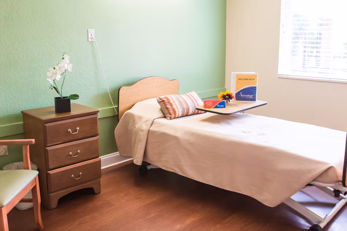 A simple bedroom in a senior living facility with a single bed covered in a beige blanket, a decorative pillow, and a small overbed table holding a welcome book and a small flower arrangement. Next to the bed is a wooden nightstand with three drawers and a white orchid plant on top. A wooden chair with a green cushion is partially visible. The walls are painted green and beige, and there is a window with blinds letting in natural light.