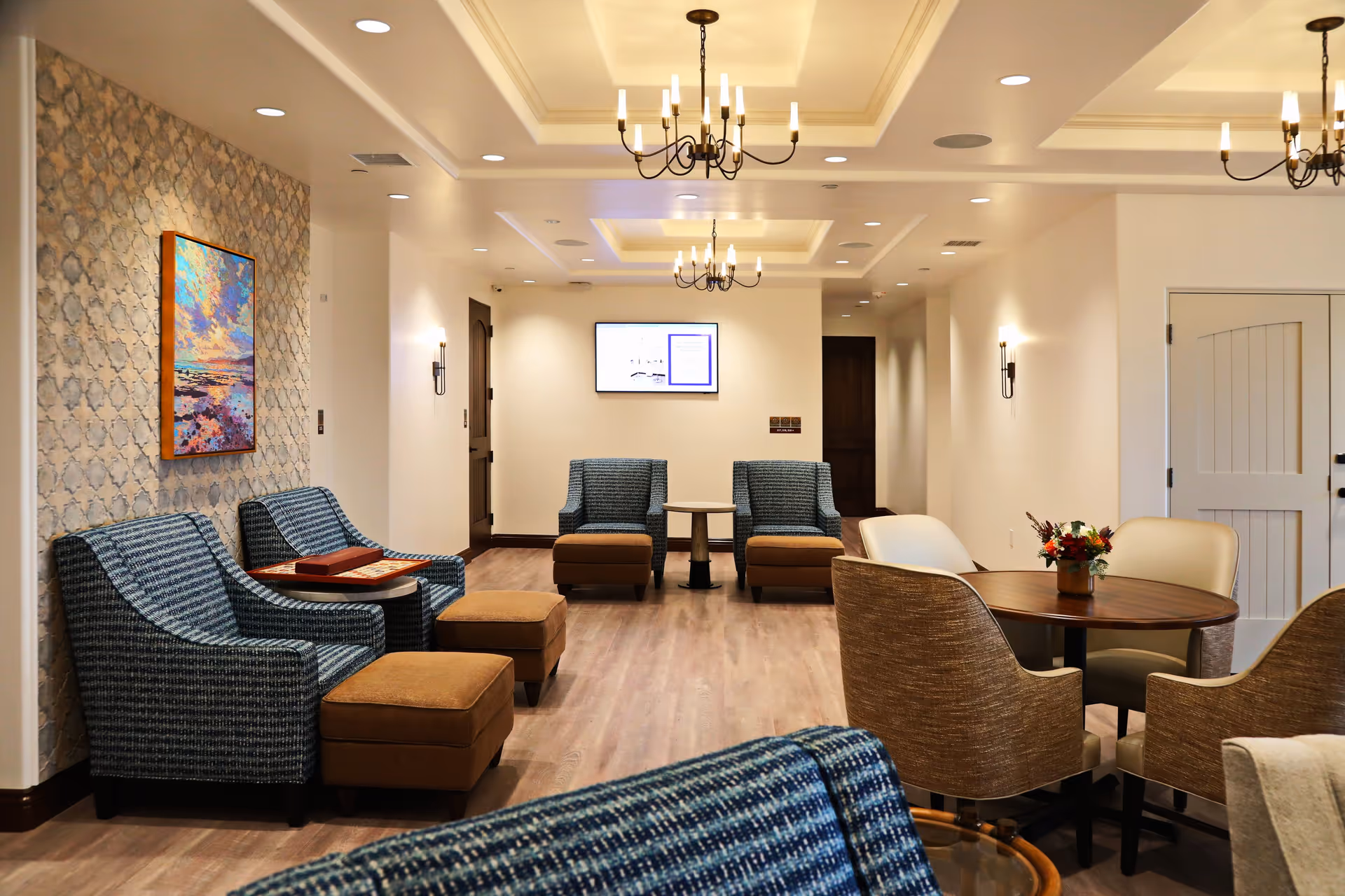 A well-lit common area with multiple seating options including blue patterned armchairs with brown ottomans, beige chairs around a round wooden table with a small flower arrangement, and a colorful painting on a textured wall. The ceiling features recessed lighting and chandeliers, and there is a digital screen mounted on the far wall.