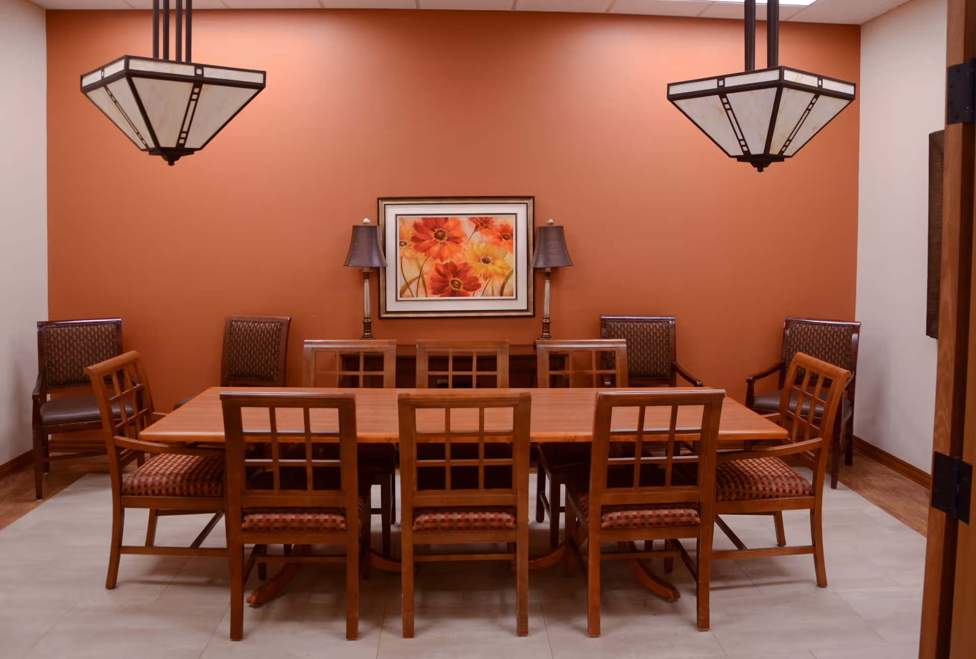 A dining room with a long wooden table surrounded by eight wooden chairs with patterned cushions. The back wall is painted orange and features a framed floral painting flanked by two table lamps. Two large pendant lights hang from the ceiling above the table.