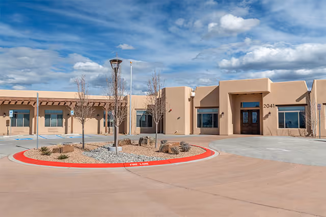 Exterior view of a single-story beige stucco building with the address number 2041. The building has several windows and a main entrance with double doors. In front of the building is a circular landscaped area with rocks, small plants, and leafless trees. The sky is partly cloudy.