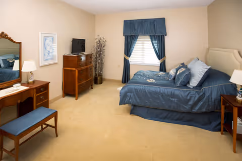 A bedroom with beige walls and carpet, featuring a bed with blue bedding and multiple pillows, a wooden dresser with a small TV on top, a wooden vanity with a mirror and a blue cushioned bench, a nightstand with a lamp, and a window with blue curtains.