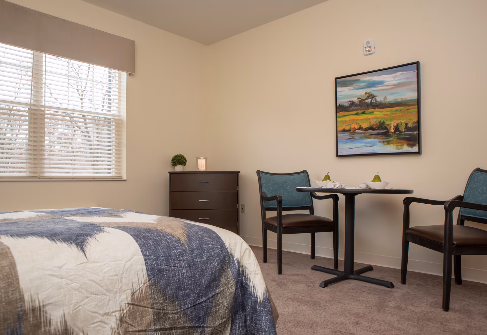 A furnished bedroom with a bed in the foreground, a dresser by the window, and a small table with two chairs under a colorful landscape painting.