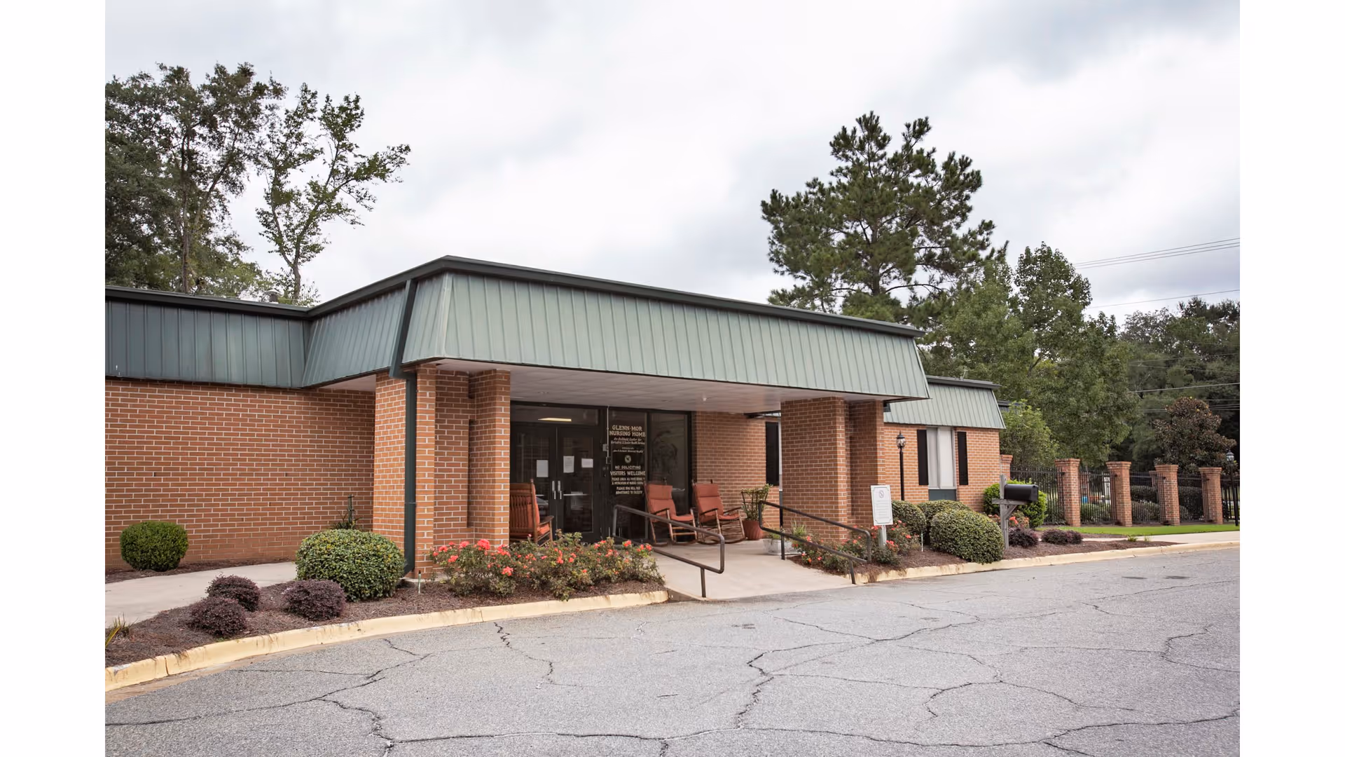 Brick senior living facility entrance with a covered portico, ramp, seating, and landscaped beds.