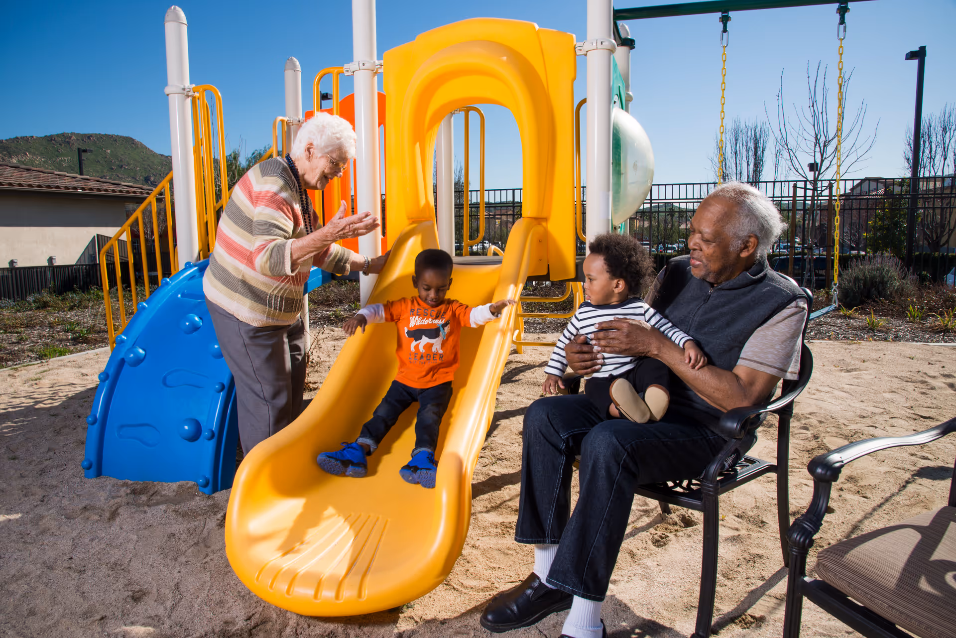 An elderly woman stands next to a yellow slide at a playground, encouraging a young boy who is sliding down. An elderly man sits nearby on a chair holding a toddler. The playground is outdoors with a fence and some buildings in the background under a clear blue sky.