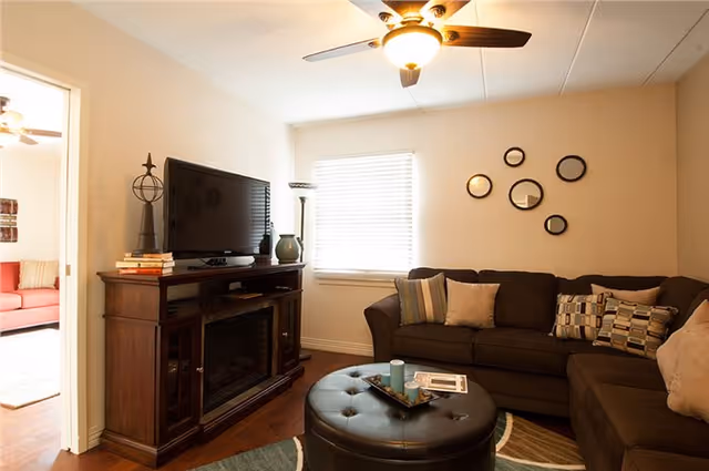 Cozy living room with a dark brown sectional sofa adorned with various patterned pillows, a round black tufted ottoman serving as a coffee table with decorative items on top, a wooden TV stand with a flat-screen television and decorative objects, a window with blinds, and a ceiling fan with a light fixture. Adjacent room with a red sofa is partially visible through an open doorway.