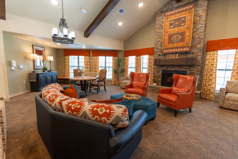 A cozy living room area in a senior living facility with a black sofa adorned with patterned cushions, two red leather armchairs, a blue ottoman, and a round wooden table with chairs near large windows with orange and yellow curtains. The room features a stone fireplace with a decorative textile hanging above it and the word 'HOME' displayed on the mantel. The ceiling has exposed wooden beams and a chandelier, and there is a sideboard with lamps and a mirror on one wall.