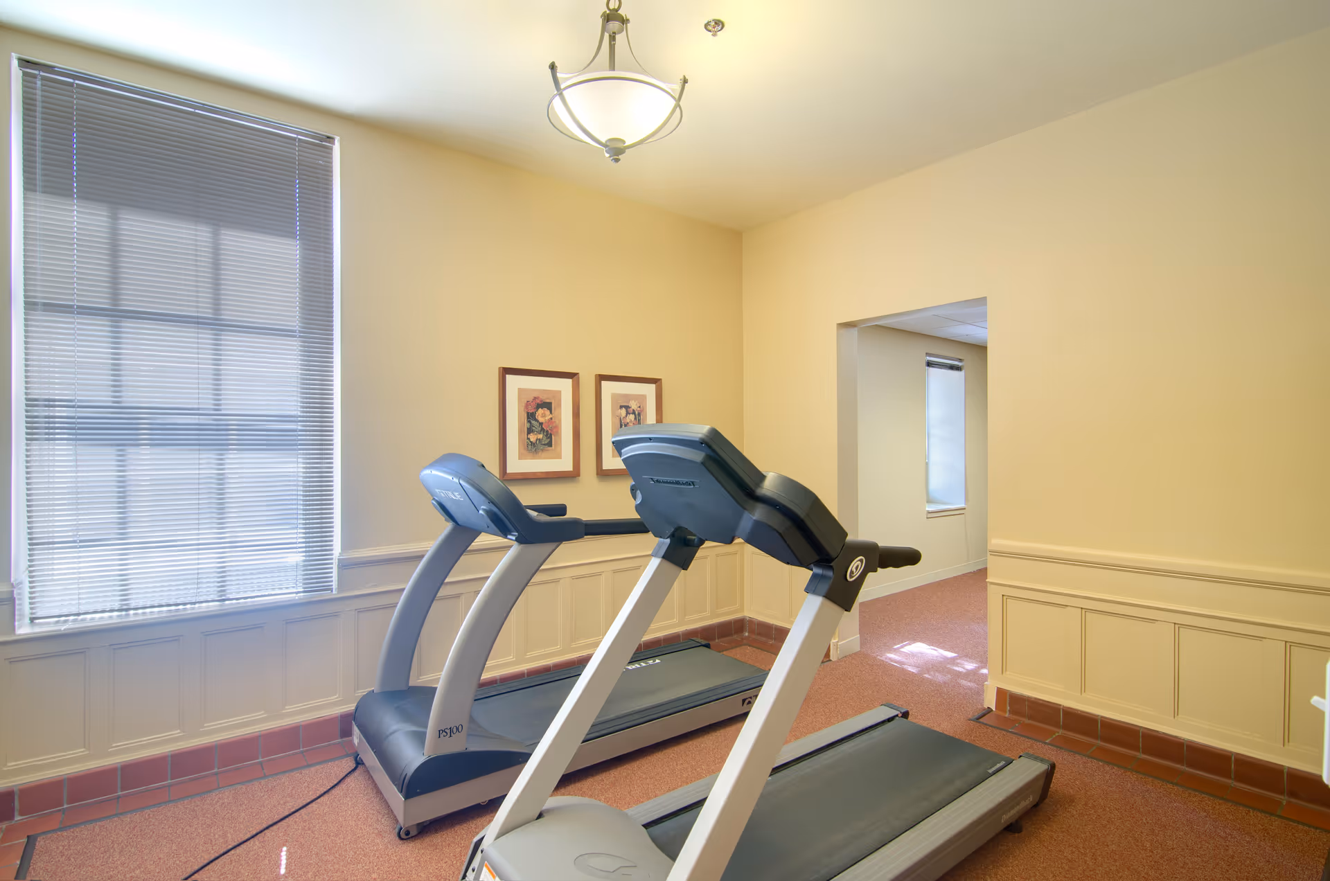 A small exercise room with two treadmills positioned side by side. The room has beige walls, a window with blinds, two framed floral pictures on the wall, and a ceiling light fixture. There is an open doorway leading to another room with a window.
