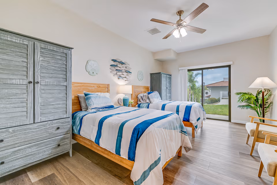 A bright bedroom with two twin beds featuring blue and white striped bedding. Between the beds is a nightstand with a lamp and decorative items. The room has wooden furniture including a wardrobe and a cabinet. A ceiling fan with lights is mounted above. There is a sliding glass door leading to a green outdoor area with palm trees and a building with a red-tiled roof. A floor lamp and two chairs with cushions are positioned near the door.