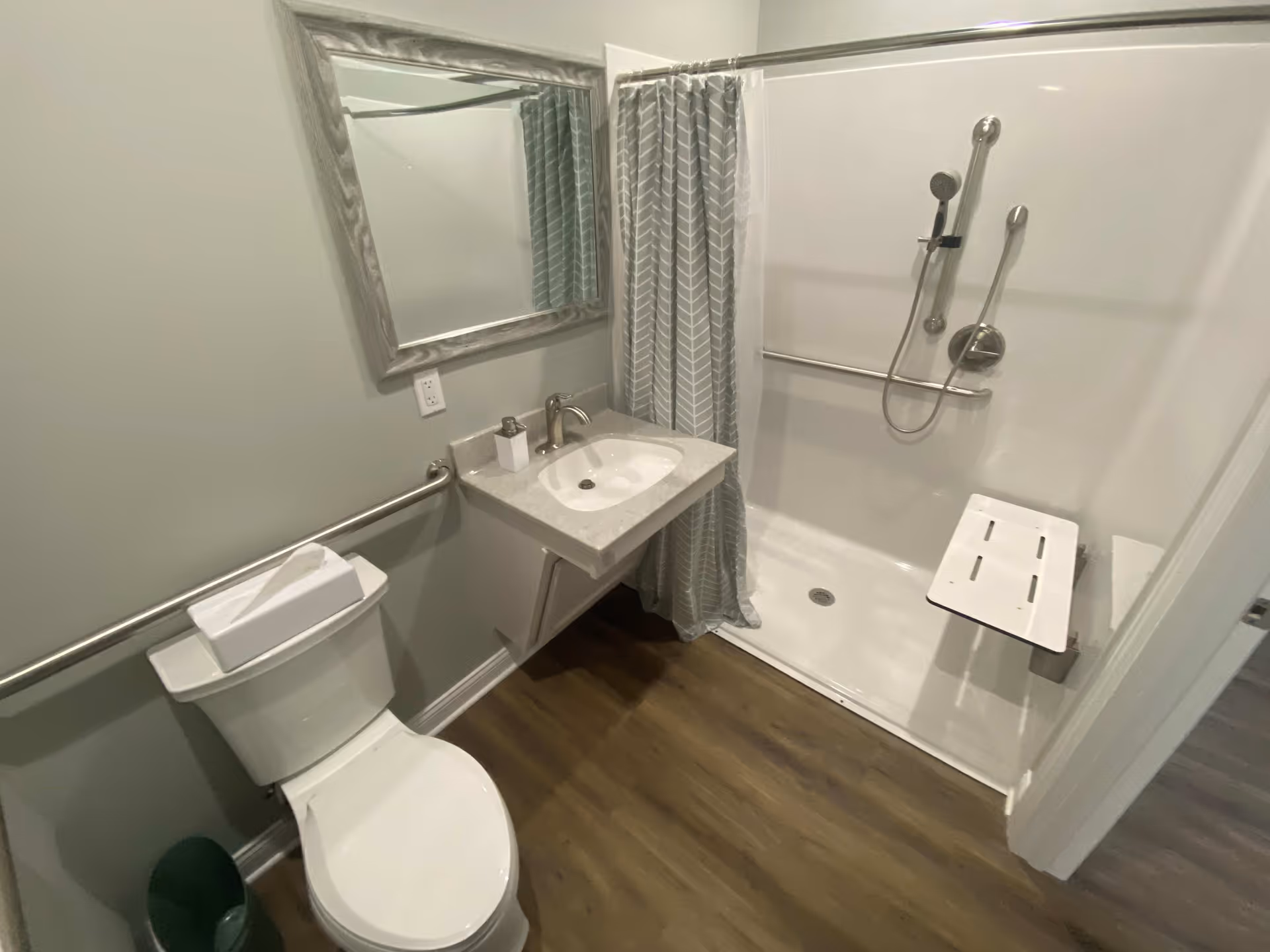 A bathroom with a toilet, a sink with a soap dispenser, a large mirror above the sink, and a walk-in shower with a shower seat and grab bars. The floor is wood-style, and the shower has a patterned curtain.