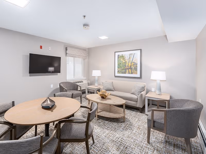 A cozy living room with a beige sofa, two gray armchairs, a round wooden dining table with four chairs, a wall-mounted flat-screen TV, two table lamps on side tables, a window with a blind, and a framed artwork on the wall.