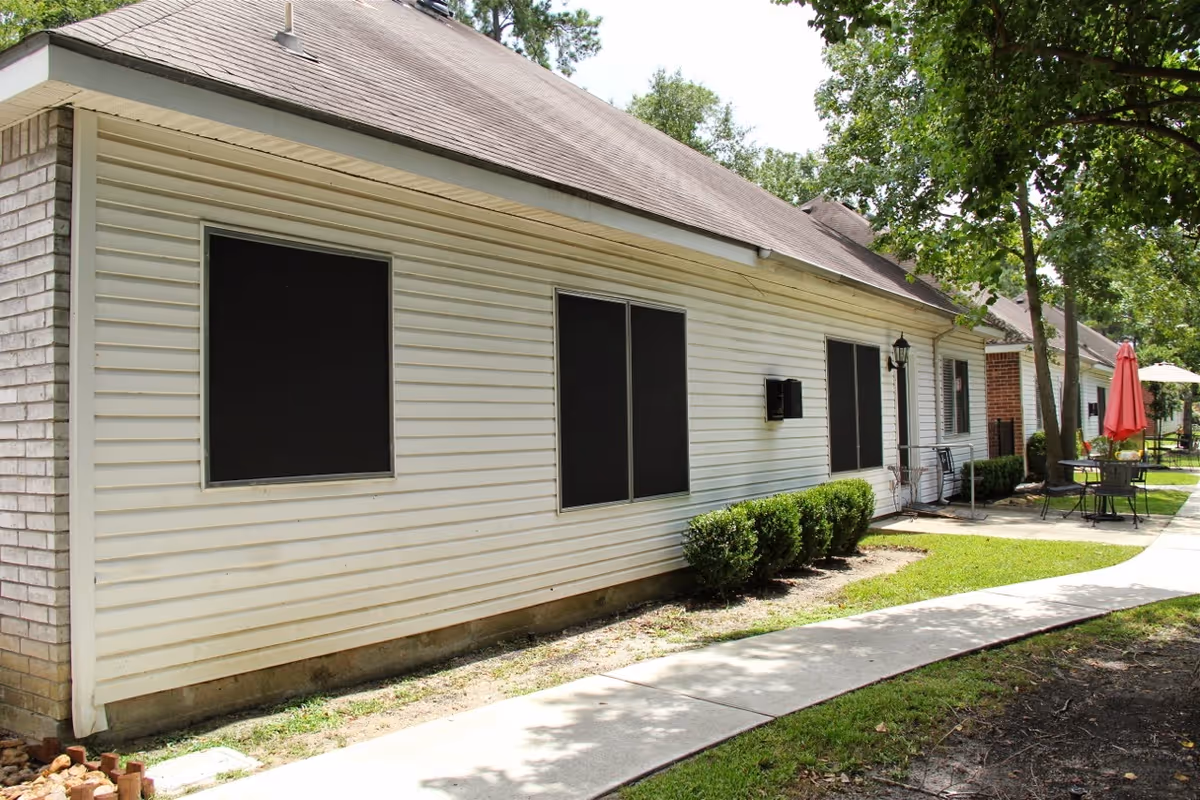 Exterior view of a single-story assisted living cottage with white siding and black window coverings. A concrete walkway runs alongside the building, bordered by small bushes and trees. Outdoor seating with tables and umbrellas is visible further down the path.