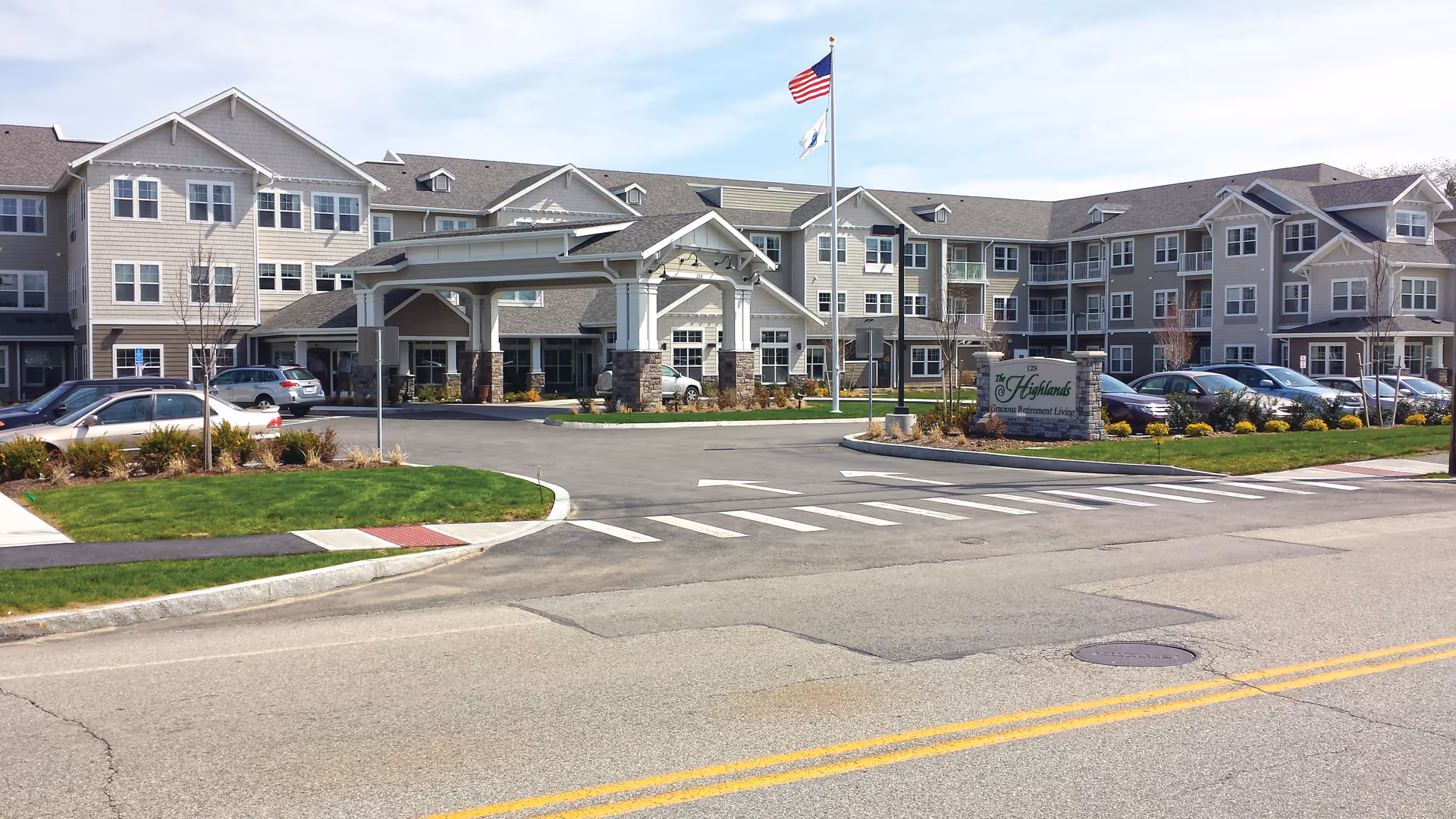 Front entrance of The Highlands Gracious Retirement Living, a multi-story senior living building with a covered porte-cochère, flagpoles, and parked cars.