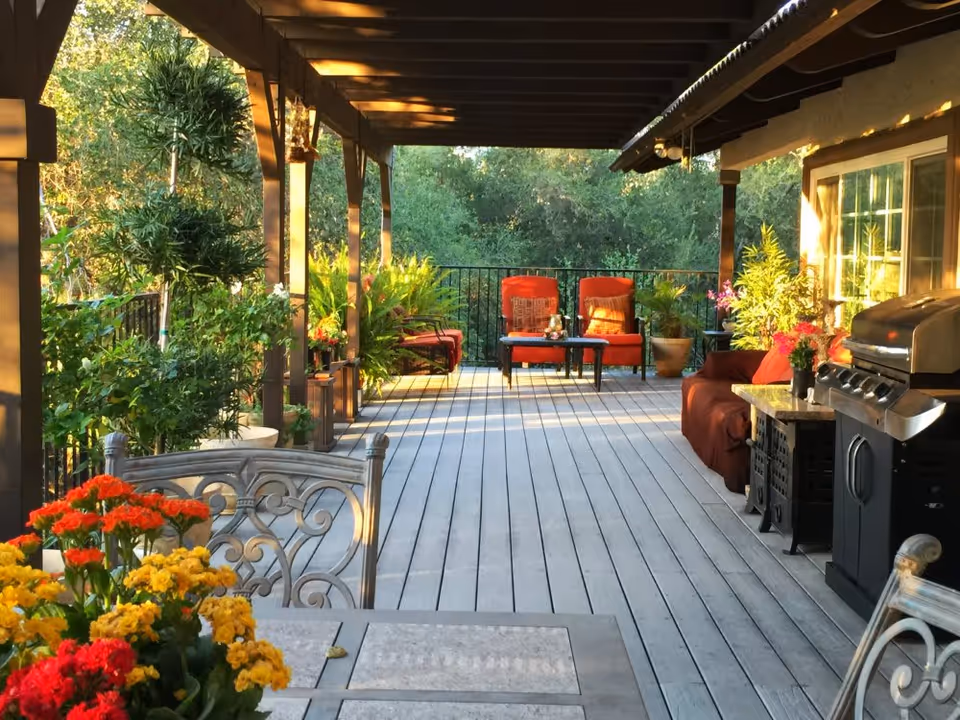 Covered wooden patio with potted plants, red cushioned chairs, a grill, and a dining table overlooking greenery.