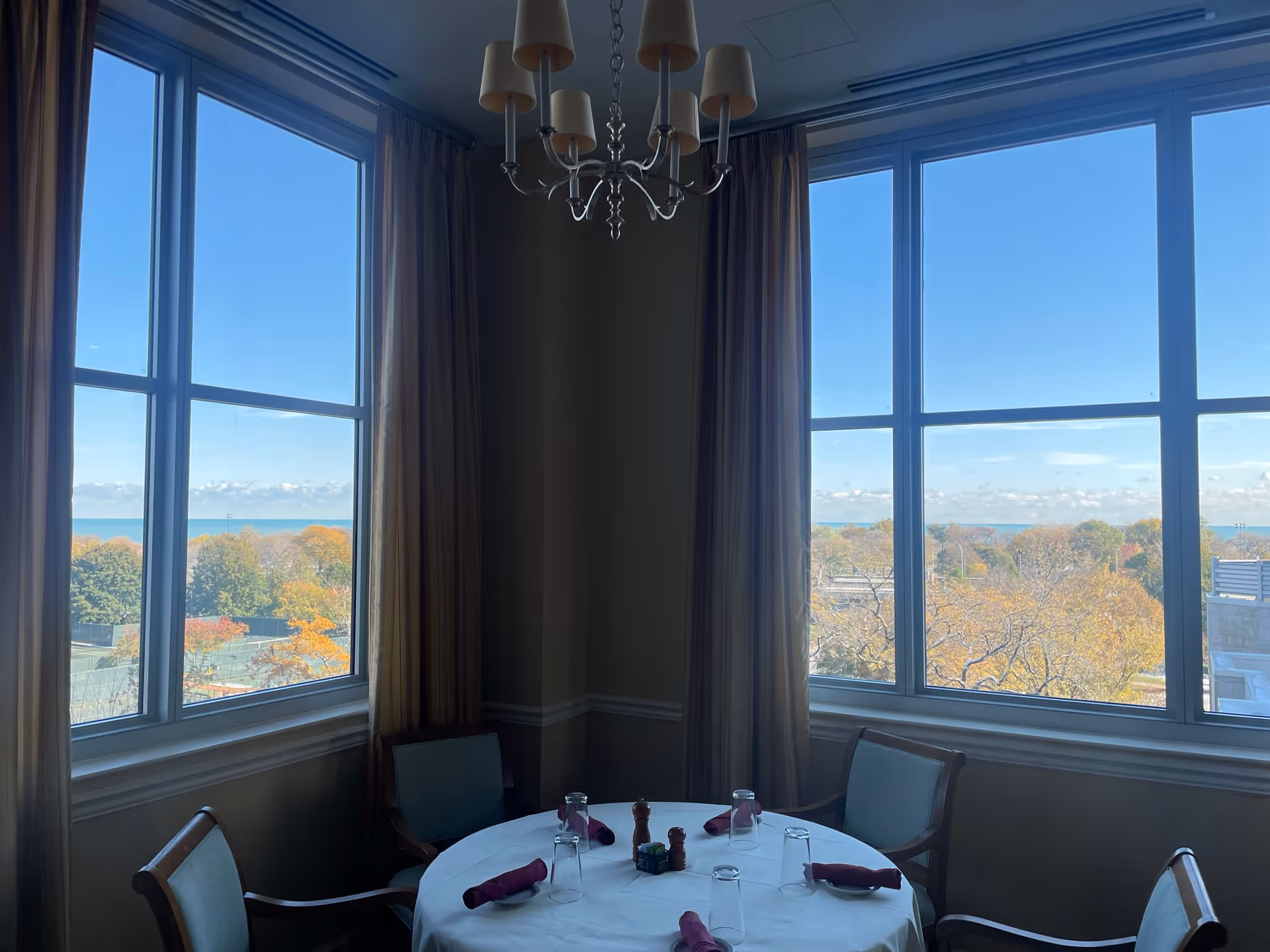 A dining room with a round table covered with a white tablecloth, set with upside-down glasses and folded burgundy napkins. Four chairs surround the table. Large windows with beige curtains offer a view of trees with autumn foliage and a clear blue sky outside. A chandelier with multiple lampshades hangs above the table.