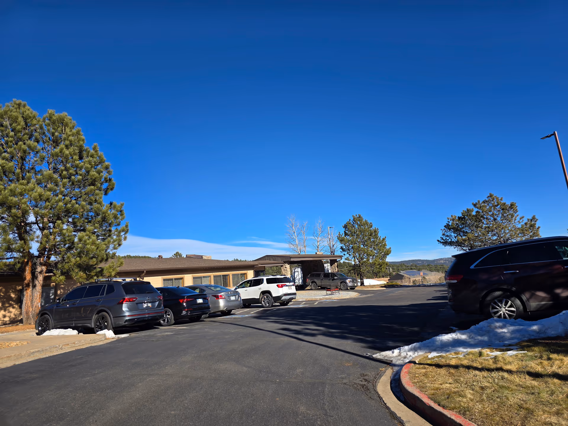 Parking lot outside a single-story building with several parked cars, surrounded by pine trees and patches of snow under a clear blue sky.