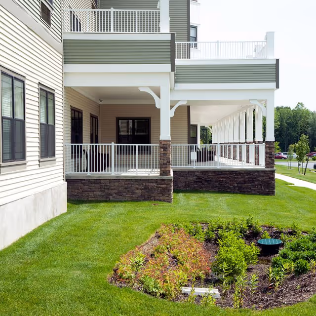 Exterior view of a senior living facility showing a covered porch with white railings and stone pillars, adjacent to a well-maintained lawn and landscaped garden bed with small plants and shrubs.