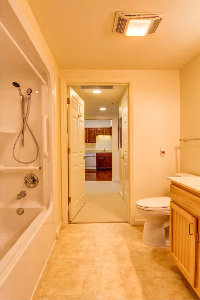 View of a bathroom with a bathtub and shower on the left, a toilet and wooden vanity with a sink on the right, and an open door leading to a hallway and kitchen area with wooden cabinets in the background.