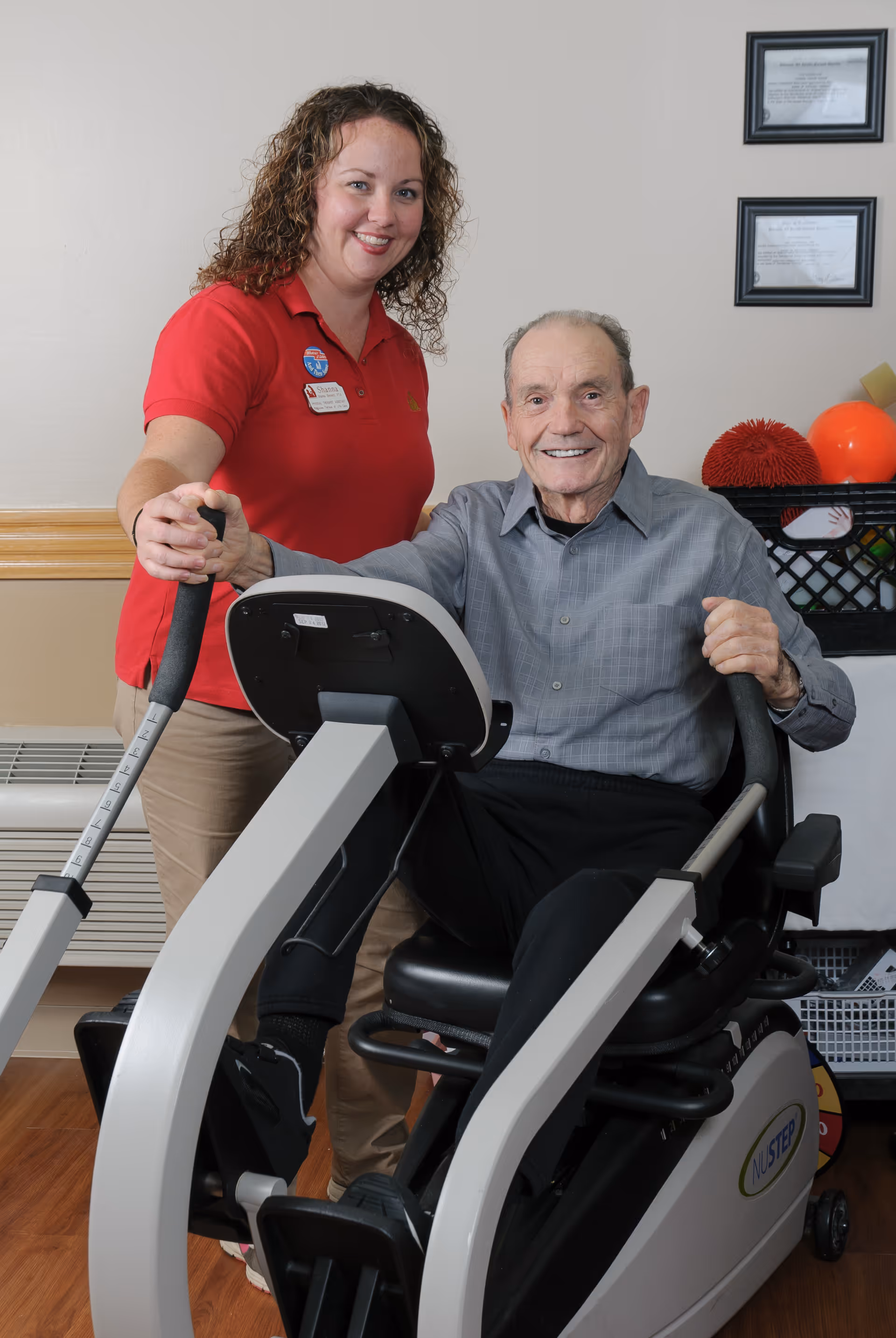 A smiling elderly man is seated on a NuStep exercise machine, holding the handles, while a female caregiver in a red polo shirt stands beside him, also holding the machine's handle. They are indoors, with framed certificates on the wall and exercise equipment in the background.