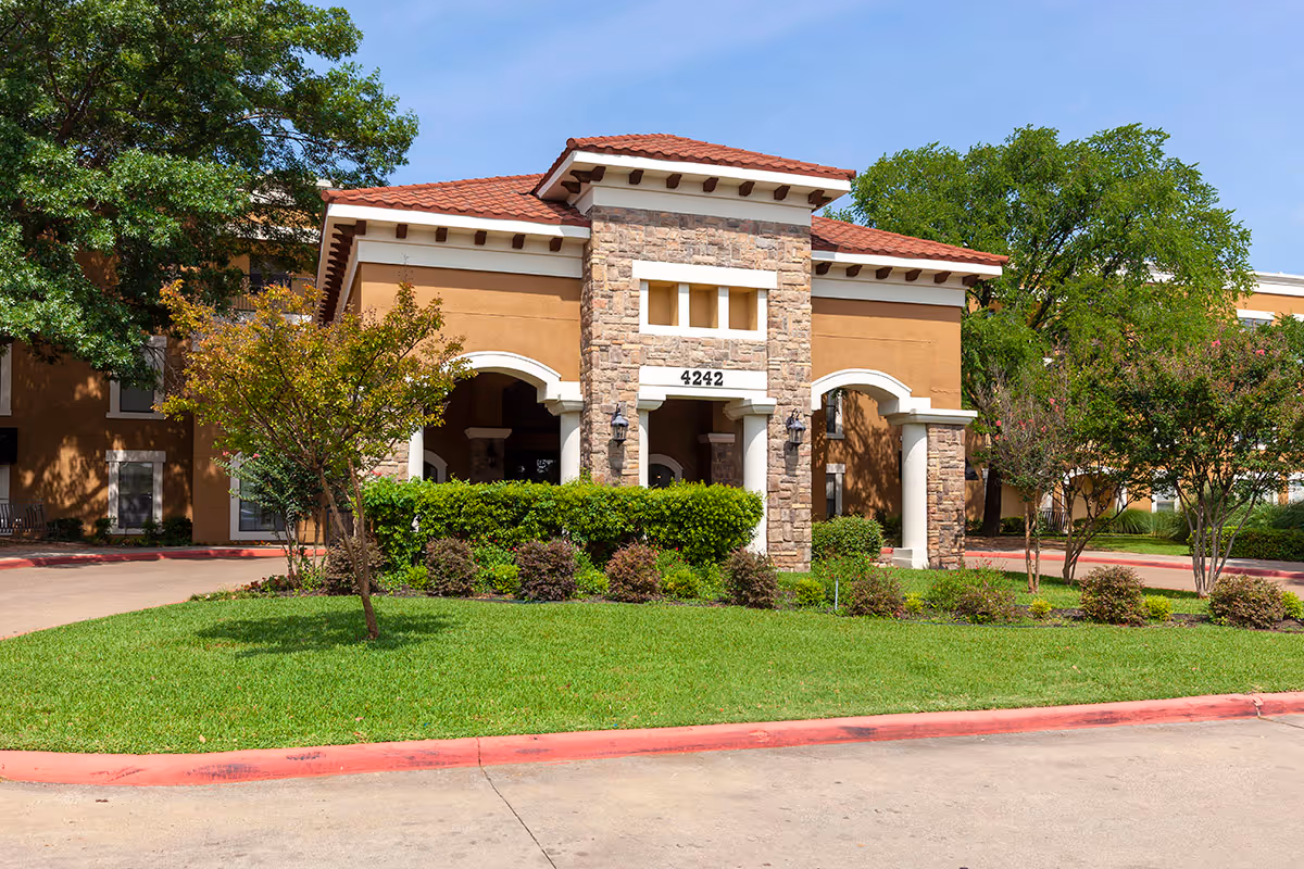 Front entrance of a two-story assisted living building with stone columns, arched entryway, and landscaped lawn showing the address 4242.
