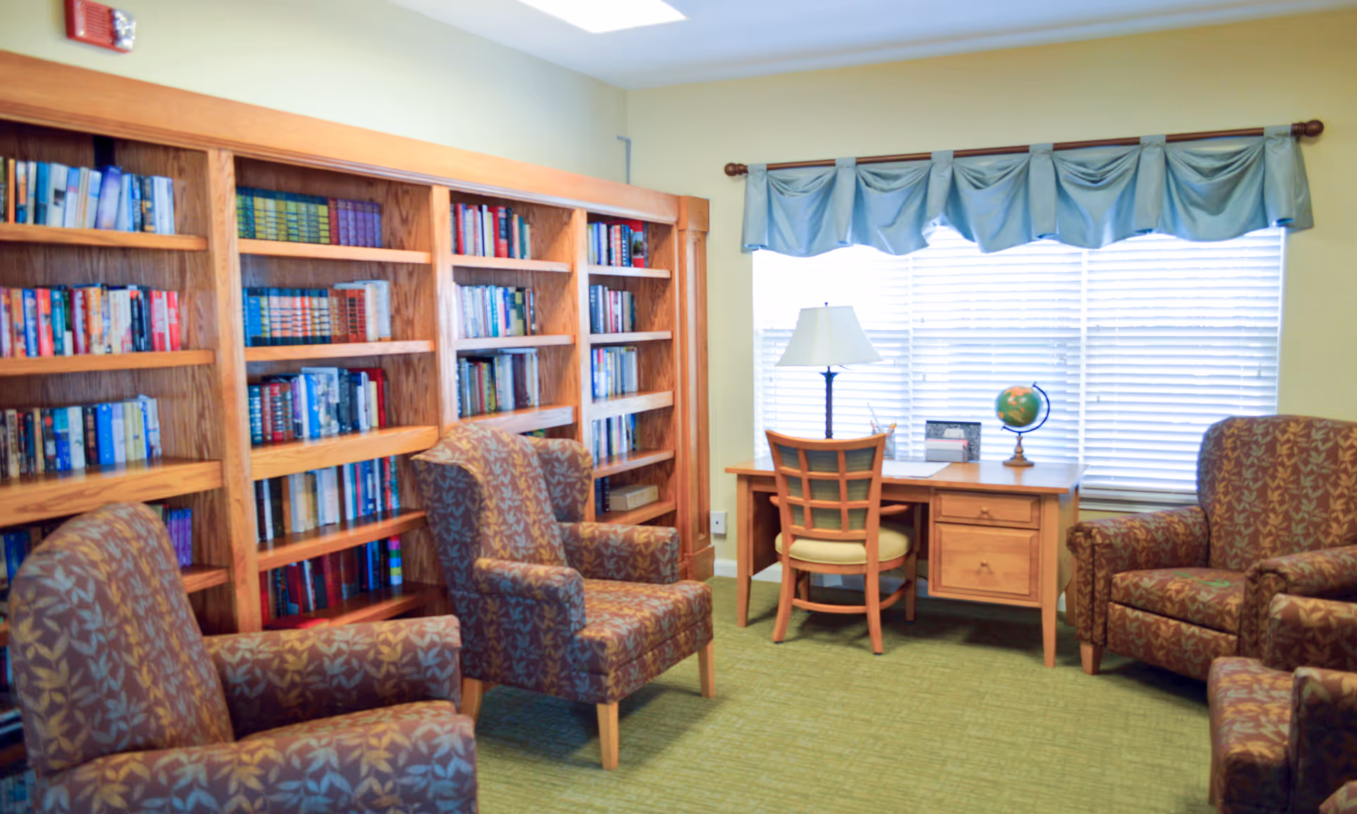 A cozy room with a wooden bookshelf filled with books, three patterned armchairs, a wooden desk with a chair, a table lamp, a globe, and a window with blinds and a blue valance.