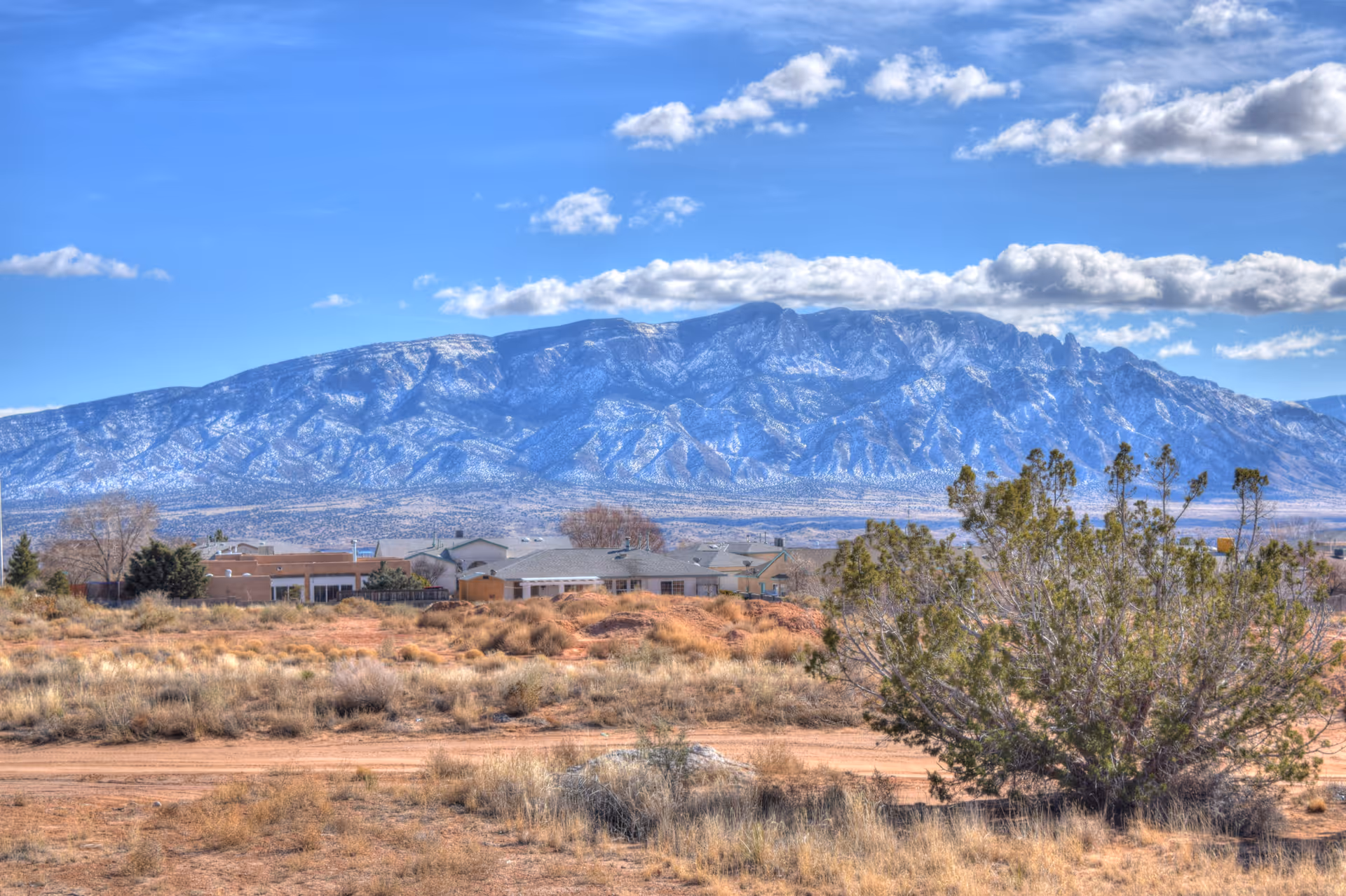 A cluster of single-story homes and scrubland with snow-dusted mountains under a blue sky in the background.