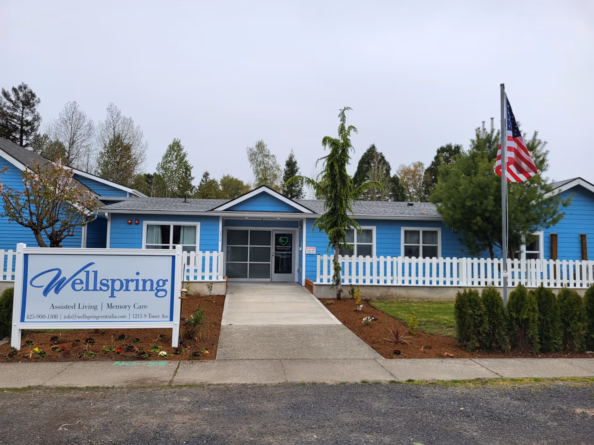 Exterior view of Wellspring Centralia, a single-story blue building with white trim and a white picket fence. There is a concrete walkway leading to the entrance, an American flag on a flagpole to the right, and a sign in front displaying the facility's name, services, contact number, email, and address. Trees and shrubs surround the building under an overcast sky.