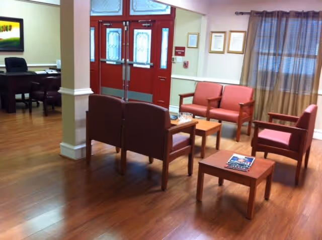 A seating area in a senior living facility with wooden floor, red cushioned chairs arranged around small wooden tables, a window with curtains, and a red double door with glass panels in the background. There is also a desk with chairs visible to the left side of the image.