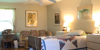 A cozy bedroom with a single bed covered with a quilt, two bedside tables with lamps, a sofa with patterned cushions, framed artwork on the walls, and a window with light curtains.