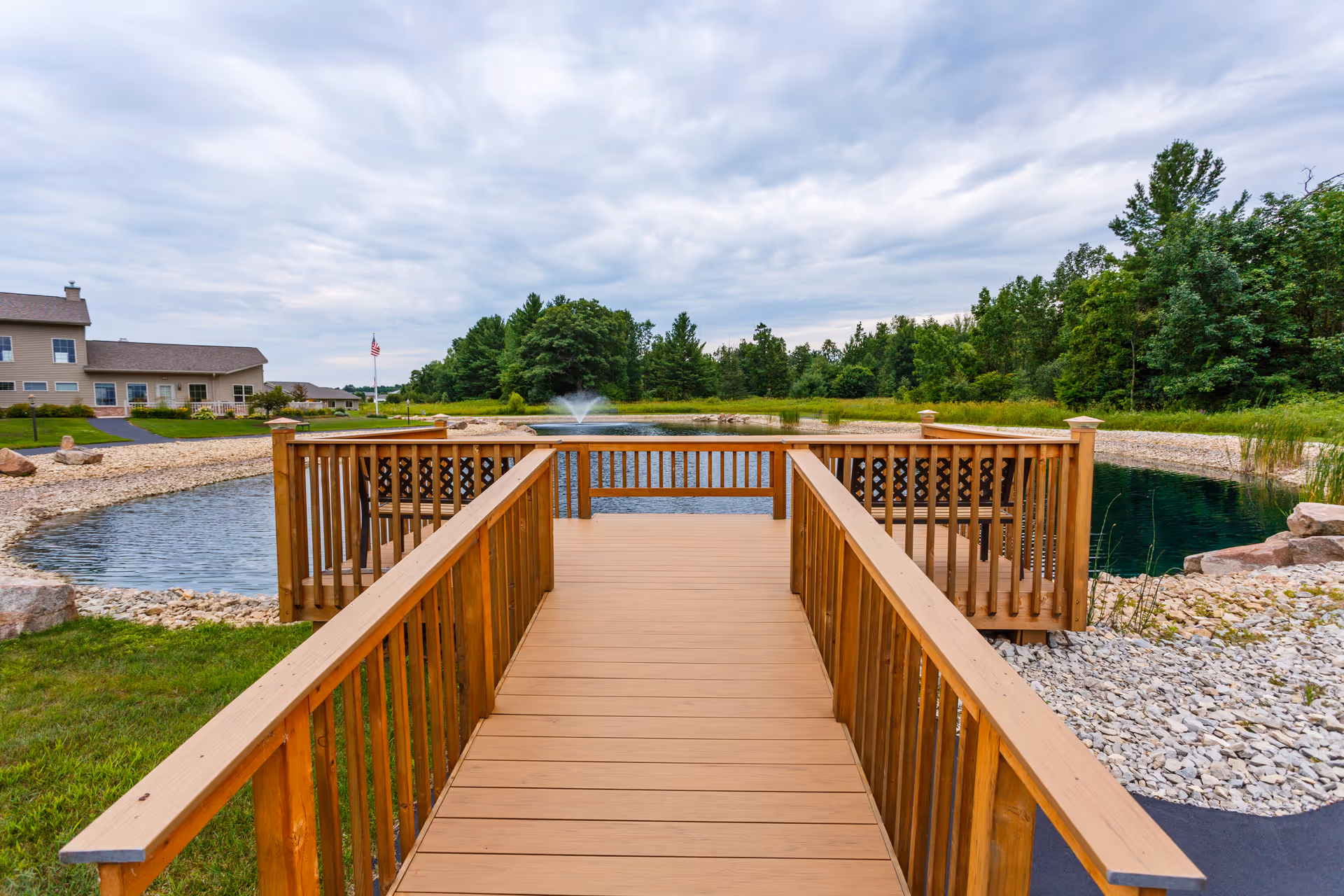A wooden walkway leading to a small deck overlooking a pond with a water fountain in the center. The pond is surrounded by rocks and greenery, with trees and a building visible in the background under a cloudy sky.