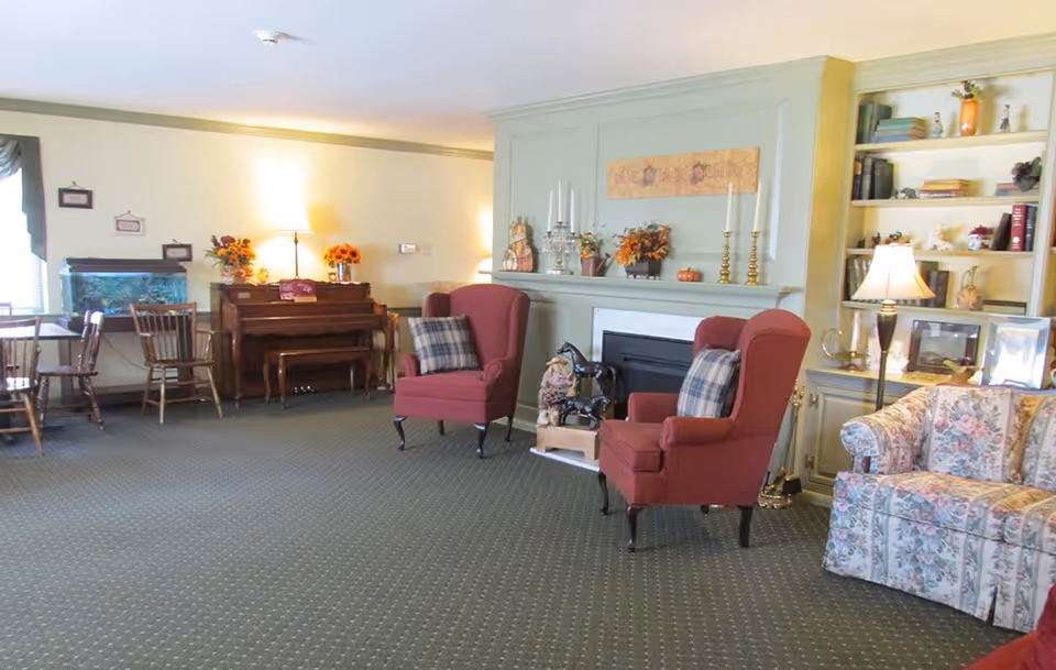 A cozy living room area in a senior living facility featuring two red armchairs with plaid pillows, a floral patterned sofa, a fireplace with decorative items on the mantel, a wooden piano with a lamp and flower arrangements on top, a fish tank, wooden chairs and tables, and built-in shelves with books and decorative objects.